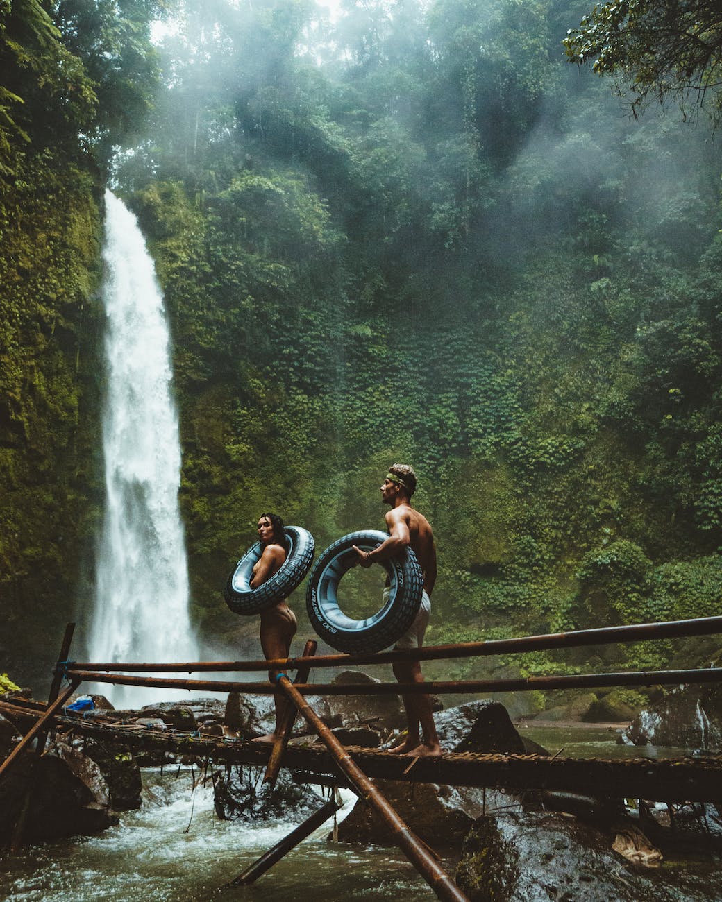 two person carrying black inflatable pool float on brown wooden bridge near waterfalls