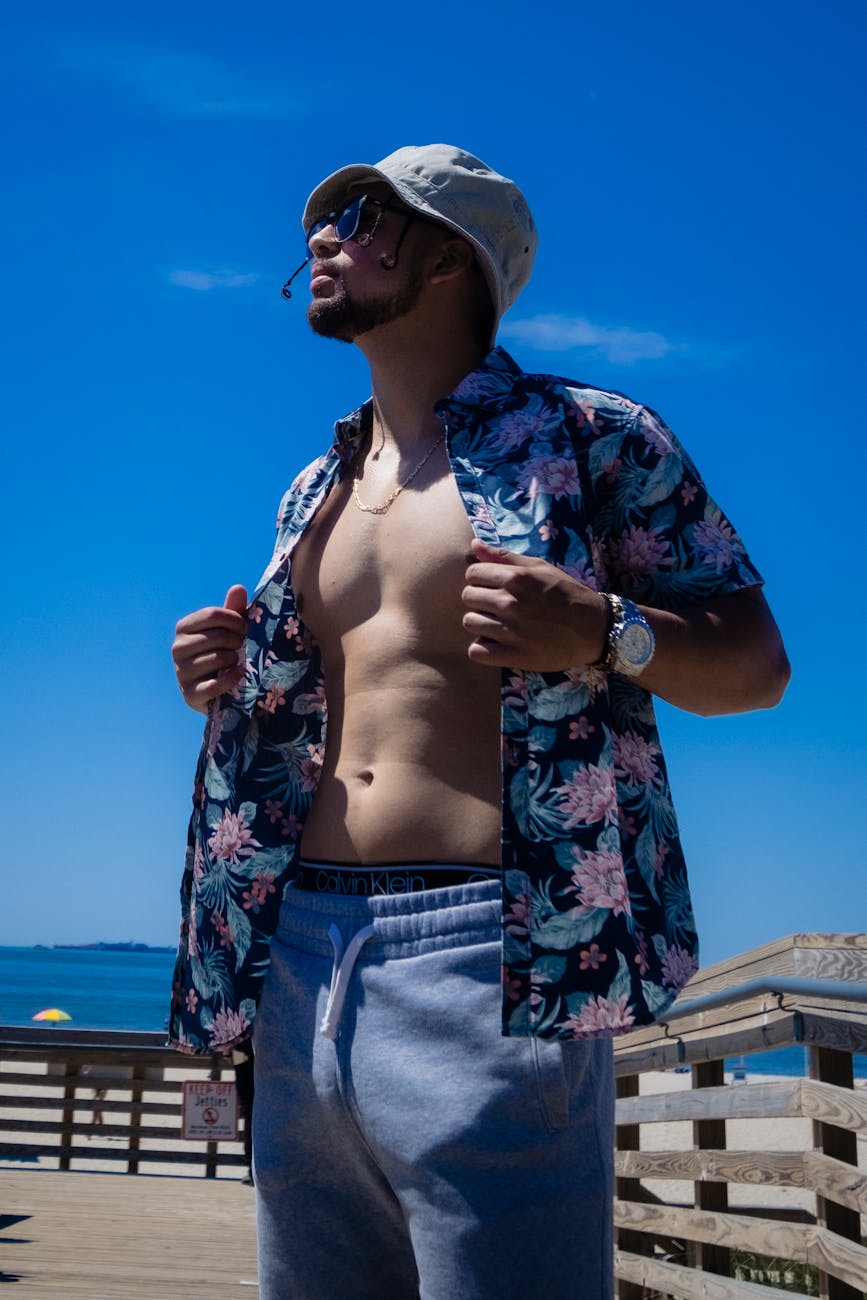 man in floral button up shirt and blue sweat shorts with bucket hat standing under blue sky